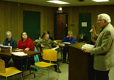 Brother John Lichi speaking from a podium with some people in a service of Cornerstone in the meeting room of the public library.